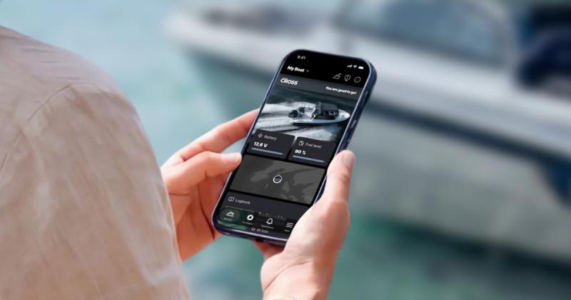 In the foreground, a smartphone in a person’s hands. The person’s upper arm and a bit of shoulder are also visible. The Cross App is displayed on the screen. A Cross boat is visible in the blurred background.