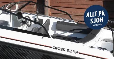 Närbild av Cross 62 V MAX:s sida och även en del av kabinen syns. I bakgrunden syns en röd timmervägg. I det övre högra hörnet finns Allt på Sjöns logotyp.