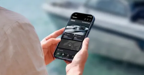 In the foreground, a smartphone in a person’s hands. The person’s upper arm and a bit of shoulder are also visible. The Cross App is displayed on the screen. A Cross boat is visible in the blurred background.